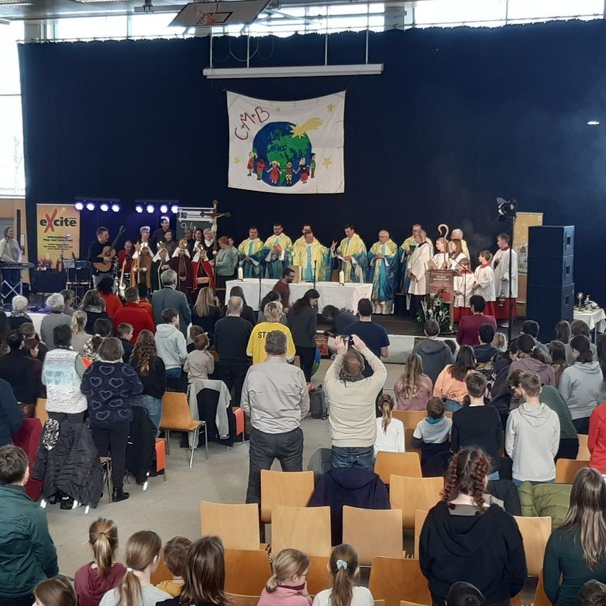 Eine Gruppe von Menschen versammelt sich in einem Saal für eine religiöse Zeremonie. Der Klerus steht auf der Bühne, während die Gemeinde in Stühlen darunter sitzt. Musikinstrumente und Banner sind auf der Bühne vorhanden.
