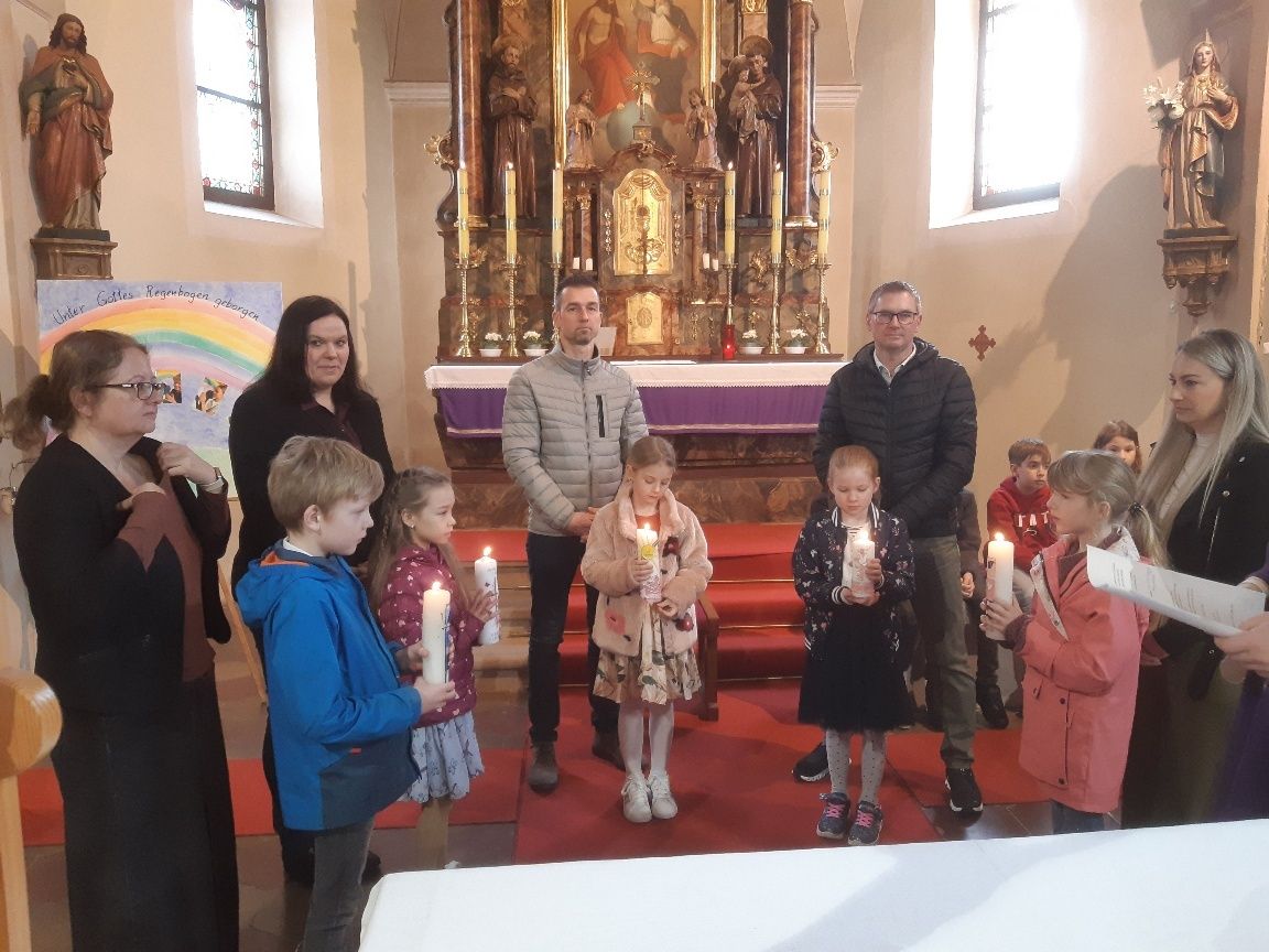 Eine Gruppe von Kindern und Erwachsenen steht in einer Kirche mit Kerzen, vor einem geschmückten Altar im Hintergrund.