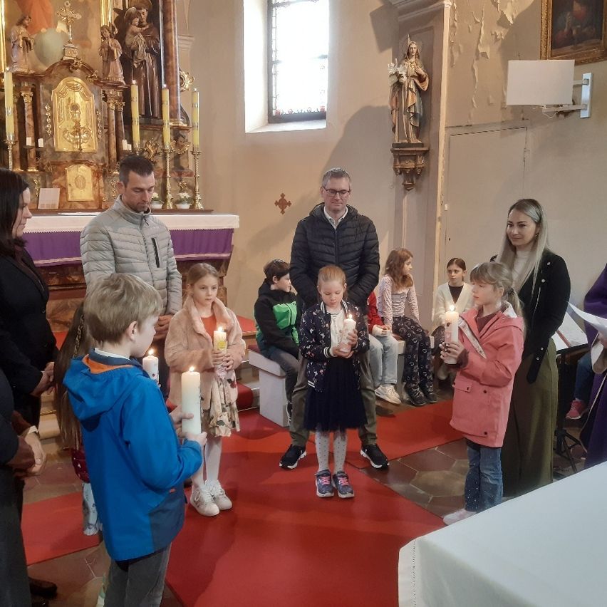Eine Gruppe von Kindern und Erwachsenen mit brennenden Kerzen steht in einer Kirche und blickt auf einen Altar mit einer Heiligenstatue und Kerzen im Hintergrund.