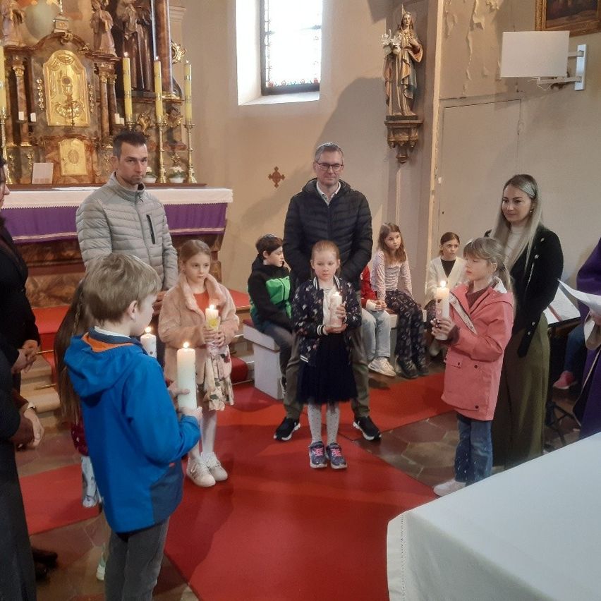 Eine Gruppe von Kindern und Erwachsenen versammelt sich in einer Kirche, hält Kerzen und ist warm gekleidet. Sie stehen auf einem roten Teppich.