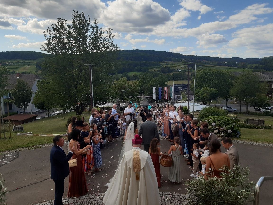 Eine Hochzeitszeremonie im Freien mit Priester, Braut, Bräutigam und Gästen unter einem blauen Himmel mit Wolken.