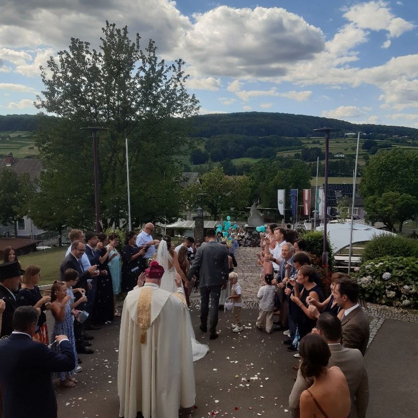 Eine Hochzeitszeremonie findet im Freien statt, wobei ein Priester die Braut und den Bräutigam anführt, umgeben von Gästen und Konfetti. Die Szenerie ist vor einem Hintergrund von Hügeln und Grünflächen angesiedelt.
