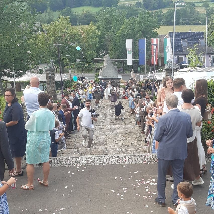 Eine Menschenmenge versammelt sich im Freien, einige beobachten einen Mann, der einen Kopfsteinpfad hinaufläuft, andere stehen an den Seiten, mit Flaggen und einer Statue im Hintergrund.