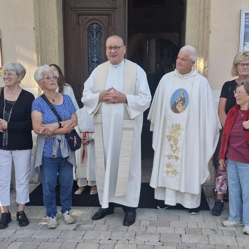 Zwei Priester stehen vor einer Kirche mit einer Gruppe älterer Frauen, alle lässig gekleidet. Die Priester tragen weiße Gewänder und einer hat ein religiöses Bild auf seinem Gewand.