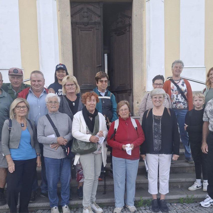 Eine Gruppe von Menschen, darunter Frauen und Männer, steht auf den Stufen eines Gebäudes, lächelt und posiert für ein Foto. Einige tragen Brillen, Hüte und Turnschuhe. Eine Frau in der Mitte hält ein Papier und einen Stift. Hinter ihnen befindet sich eine Holztür und eine weiße Wand.