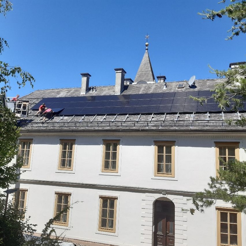 Drei Arbeiter installieren Solarmodule auf dem Dach eines weißen Gebäudes mit mehreren Fenstern und einer braunen Tür.