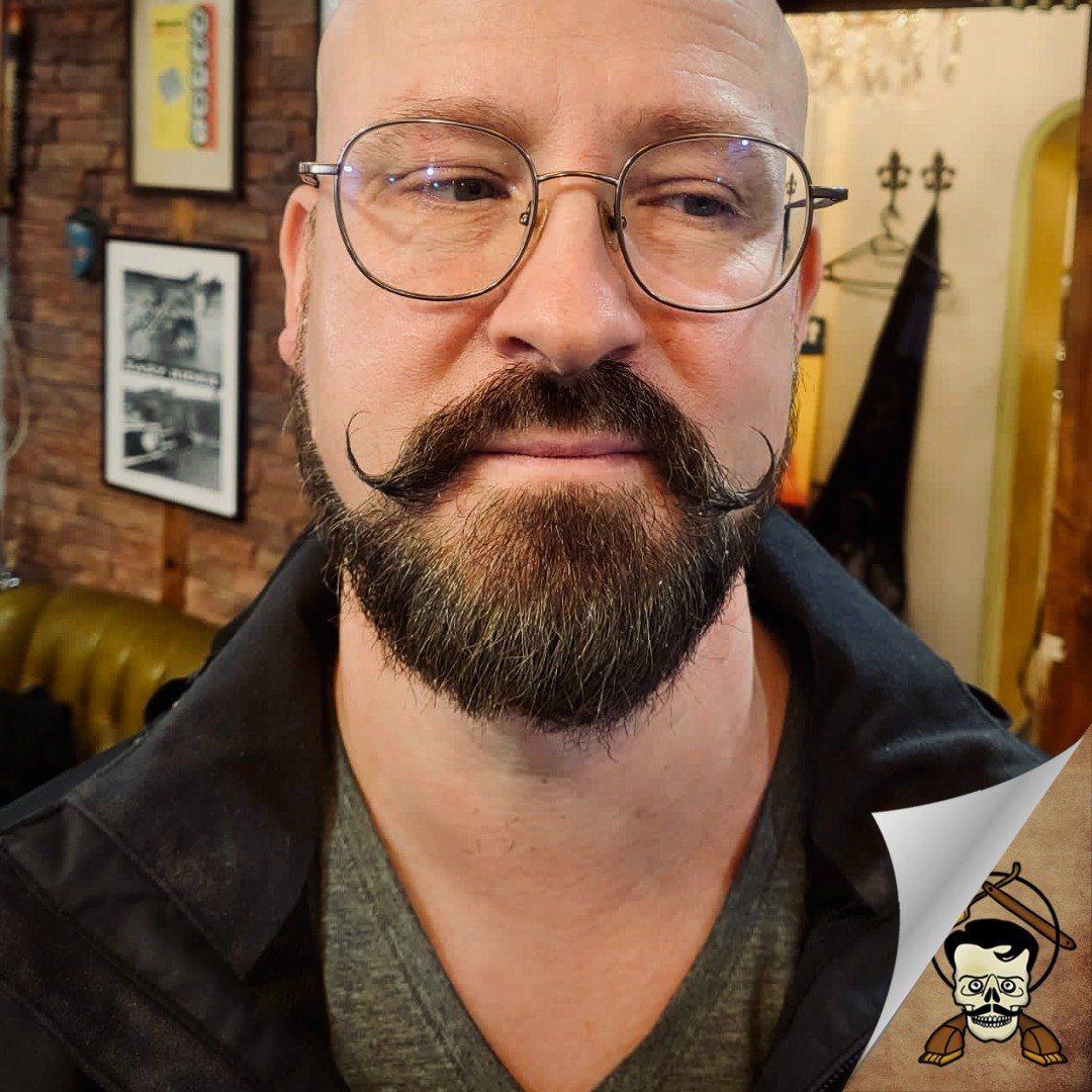 Ein Mann mit Brille, vollem Bart und Schnurrbart, vor einer Ziegelwand mit Bilderrahmen und einem schwarzen Mantel mit einem Totenkopf-Tattoo und Knochen.