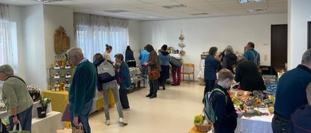 Im Inneren durchstöbern mehrere Personen verschiedene Gegenstände auf Tischen. Einige tragen Rucksäcke, und es sind Artikel wie Flaschen und Körbe ausgestellt. Der Raum ist mit Deckenleuchten beleuchtet.