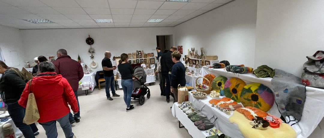 Ein Raum mit weißen Wänden und Decken zeigt verschiedene Gegenstände auf Tischen und Regalen. Menschen gehen herum, einer schiebt einen Kinderwagen.