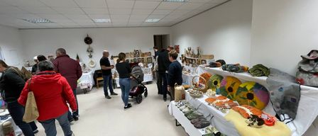 Ein Raum mit weißen Wänden und Decken zeigt verschiedene Gegenstände auf Tischen und Regalen. Menschen gehen herum, einer schiebt einen Kinderwagen.