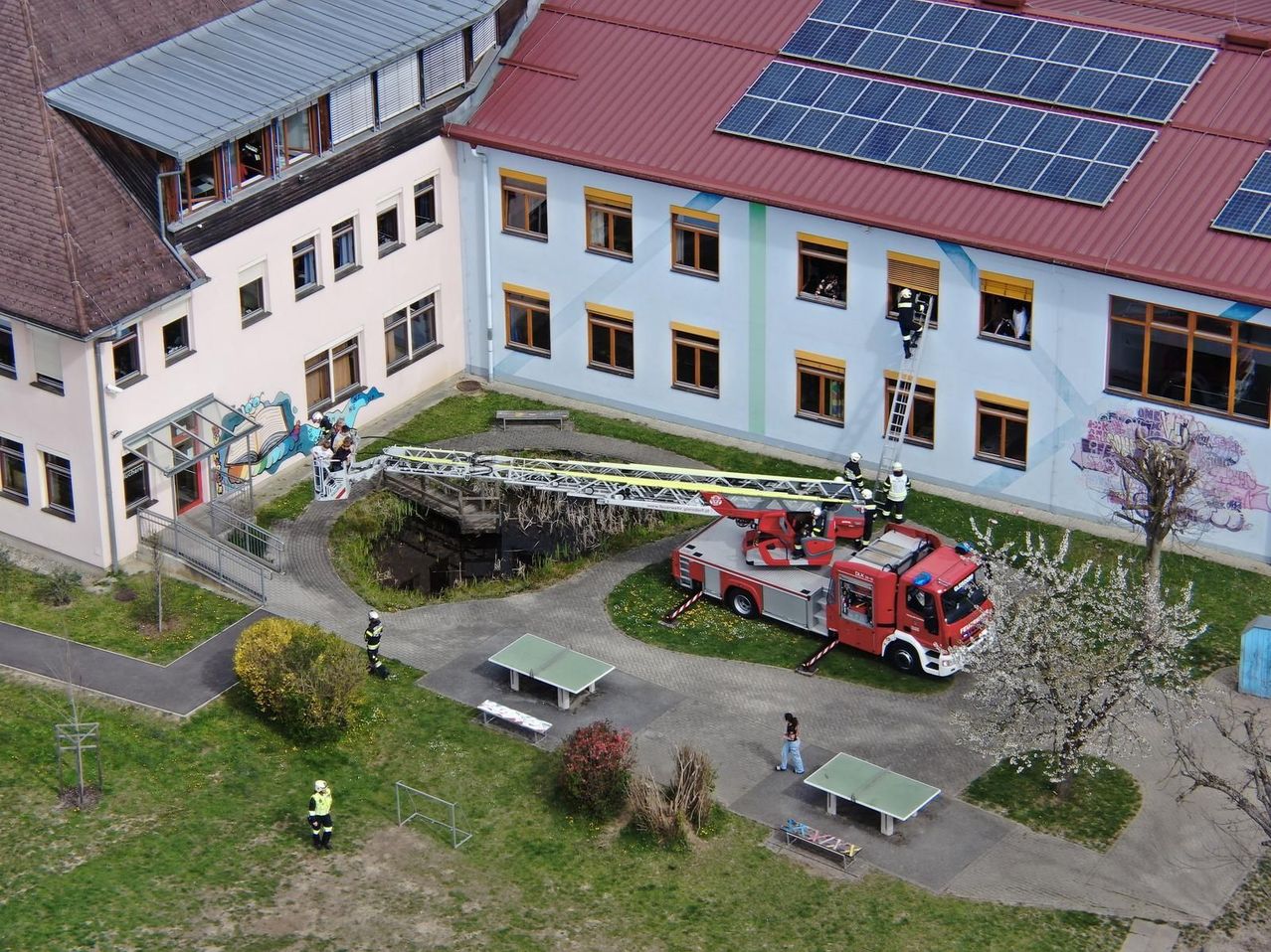 Ein Feuerwehrauto steht vor einem Gebäude mit Solarpanelen auf dem Dach. Eine Leiter ist ausgeklappt und Feuerwehrleute klettern daran hoch.