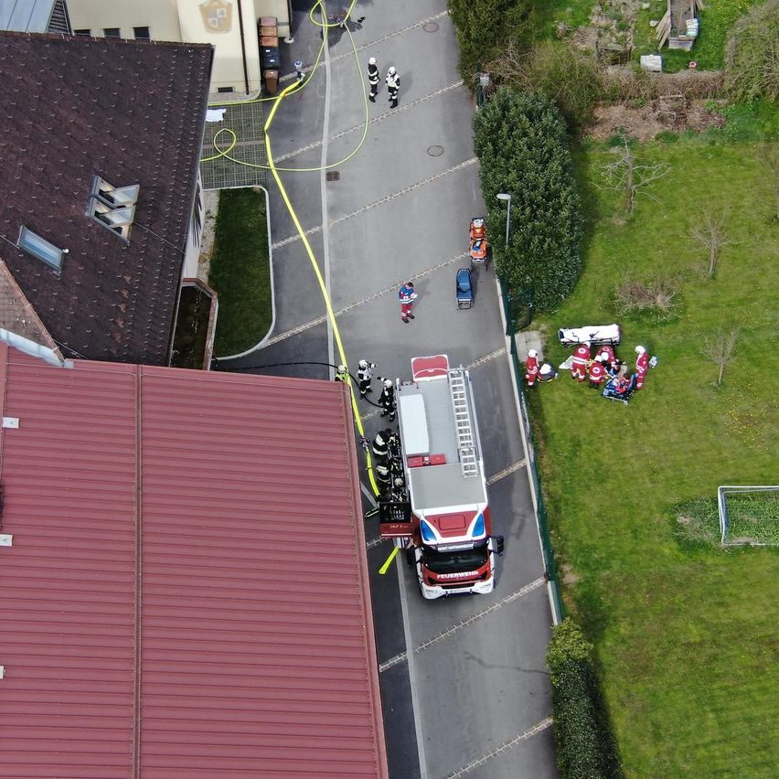 Ein Luftbild einer Notfallszene, ein Feuerwehrauto ist auf der Straße geparkt, Feuerwehrleute kümmern sich um Personen am Straßenrand, und mehrere Personen stehen herum.