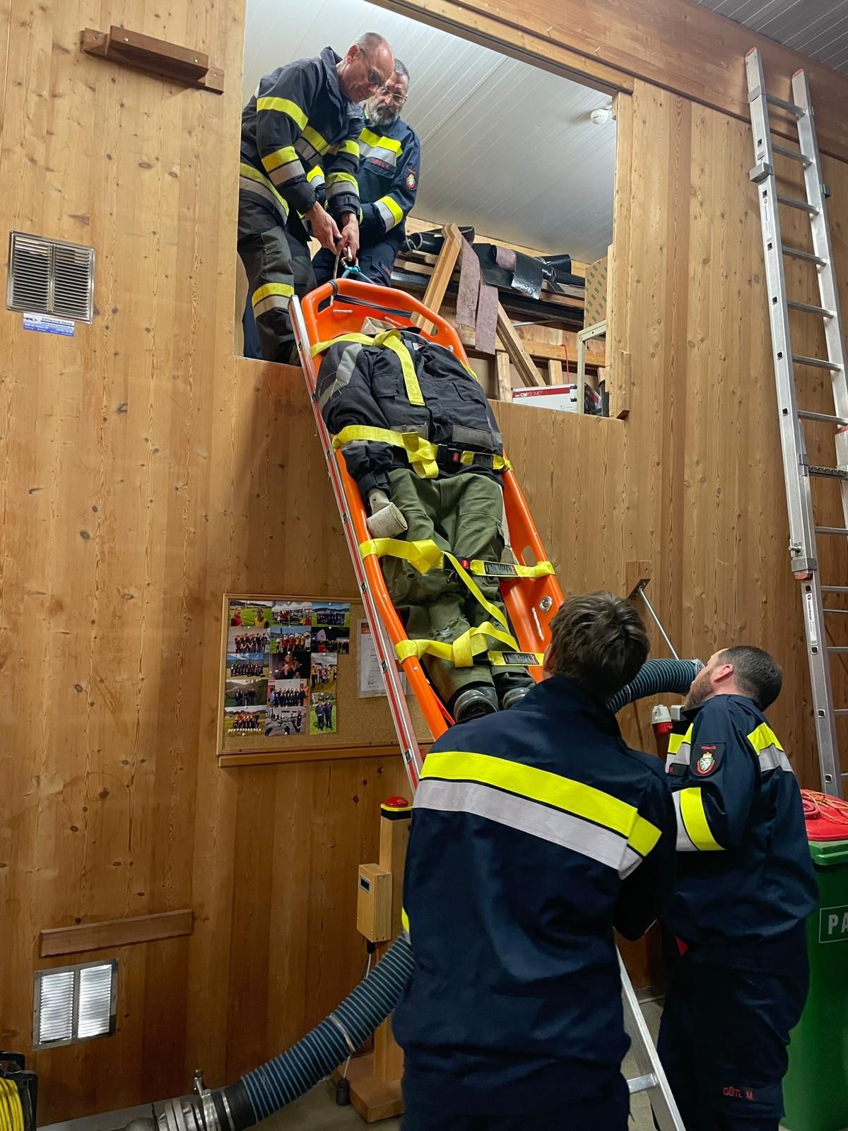 Drei Feuerwehrleute in Uniform tragen eine Person auf einer Trage durch einen Raum mit Holzwänden.