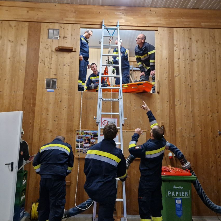 Feuerwehrleute üben eine Rettungsaktion in einem Raum mit einer Holzwand. Ein Mann steht auf einer Leiter, während ein anderer Mann zur Decke zeigt.