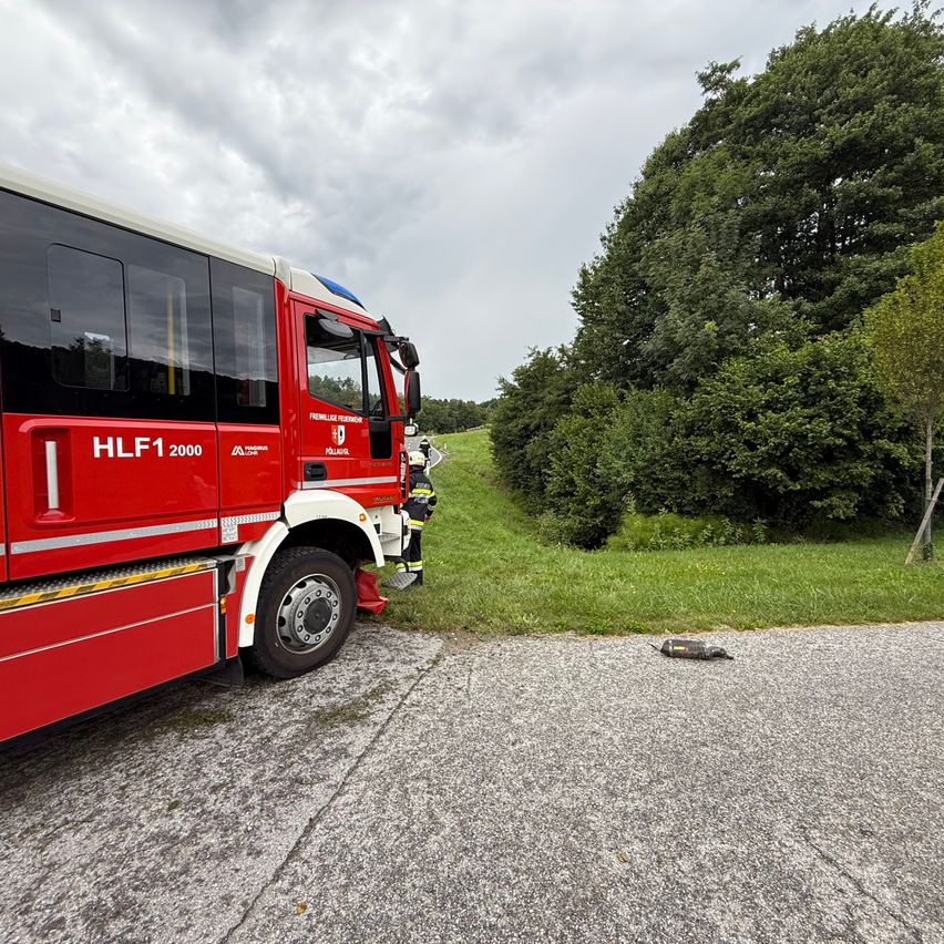 Ein roter Feuerwehrwagen mit der Aufschrift HLF1 2000 steht am Straßenrand. Ein Feuerwehrmann steht in der Nähe und ein Bohrer liegt auf dem Boden.