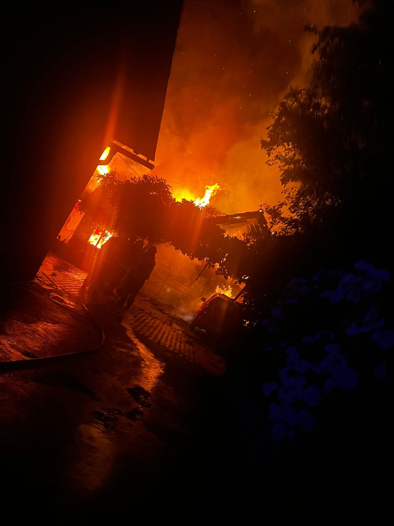 Eine Nachtszene eines brennenden Hauses mit Feuerwehrleuten davor, beleuchtet vom Feuerschein, mit aufsteigendem Rauch und Bäumen im Hintergrund.