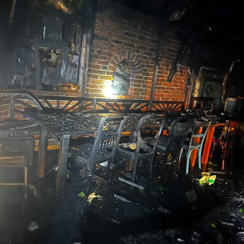 Das Innere eines abgebrannten Restaurants zeigt beschädigte Tische und Stühle, eine mit Ruß bedeckte Ziegelmauer und eine helle Lichtquelle rechts. Schutt liegt auf dem Boden verstreut.