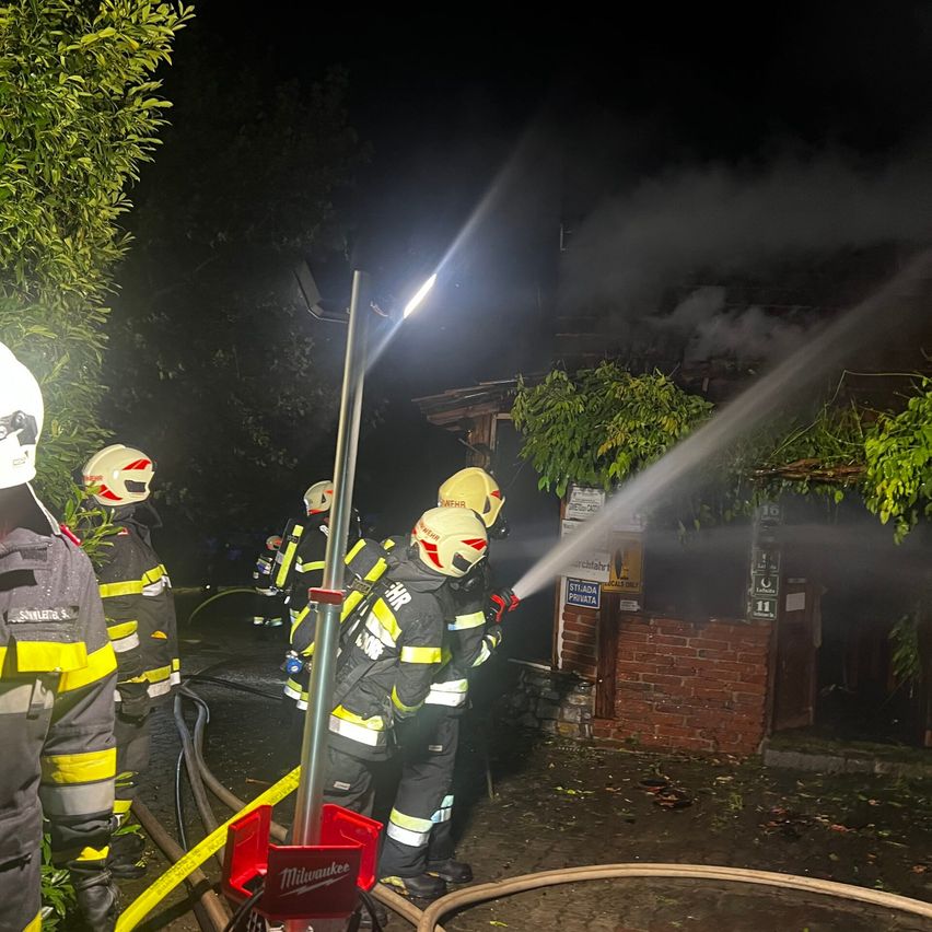Feuerwehrleute bekämpfen in der Nacht ein Feuer. Sie verwenden einen Schlauch, um Wasser auf das Gebäude zu sprühen. Die Szene ist von einem hellen Licht beleuchtet.