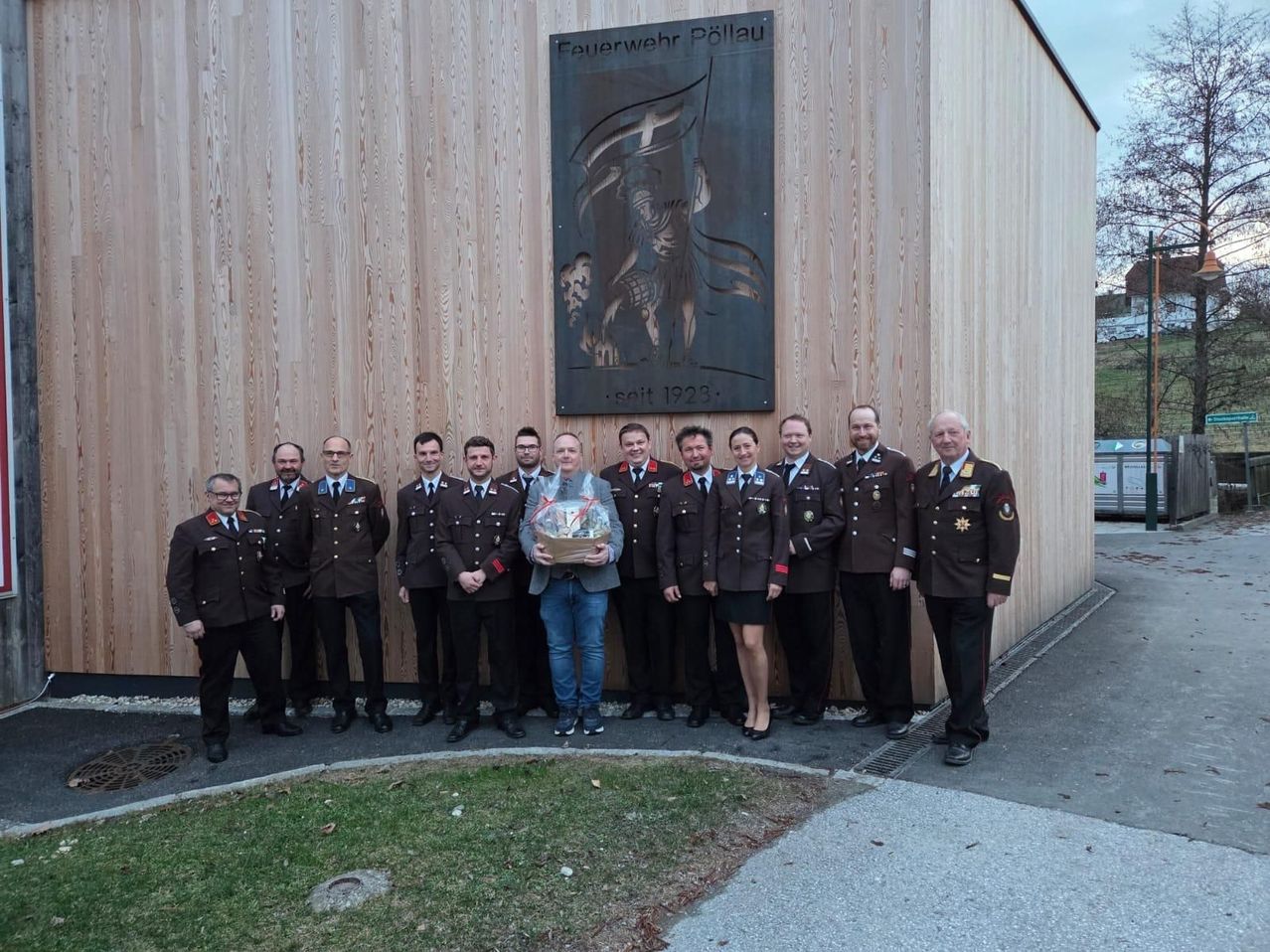 Eine Gruppe von Militärangehörigen in Uniform steht vor einem Holzgebäude, einer hält ein Geschenk. Über ihnen befindet sich eine Tafel mit einem Kreuz und dem Text 'Feuerwehr Pollau seit 1923'.