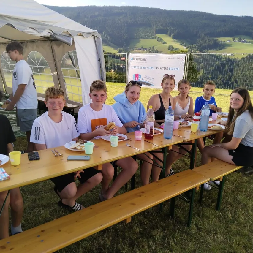 Eine Gruppe junger Menschen sitzt an einem Picknicktisch in einem Grasbereich, mit Bergen im Hintergrund. Sie genießen ihr Essen, und einige tragen Sonnenbrillen.