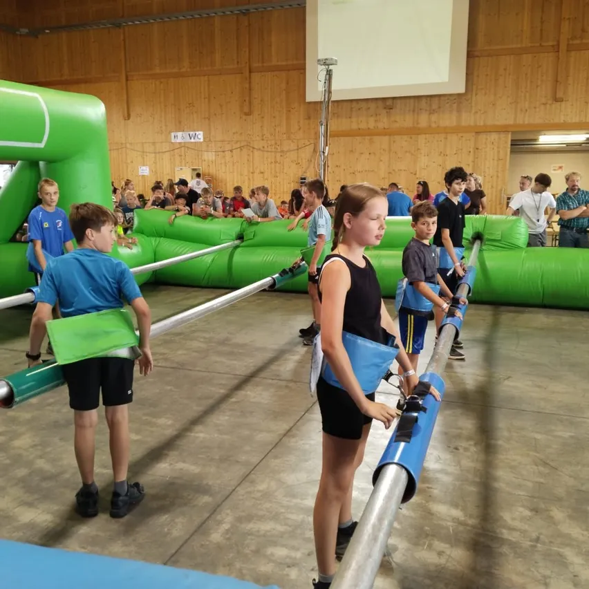 Eine Gruppe von Kindern nimmt an einem Indoor-Hindernisparcours teil, wobei sie sich an mit blauer Polsterung versehenen Stangen festhalten. Ein Mädchen in einem schwarzen Tanktop schaut nach unten.