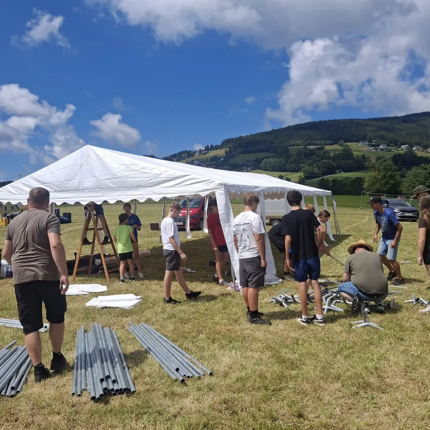 Eine Gruppe von Menschen baut ein weißes Zelt auf einem Grasfeld mit Bergen im Hintergrund auf. Einige stehen, während andere am Zelt arbeiten.