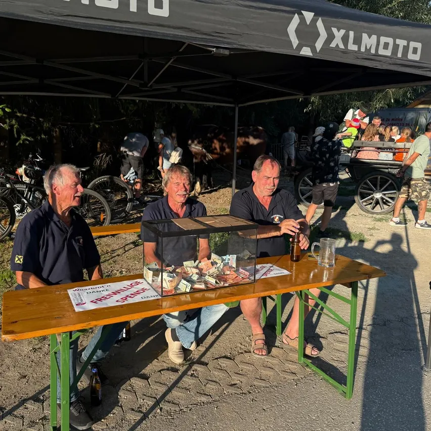 Drei Männer sitzen unter einem XLMOTO-Zelt, einer hält ein Bier und ein anderer hat eine Kiste mit Geld auf dem Tisch. Fahrräder und eine Pferdekutsche sind im Hintergrund.