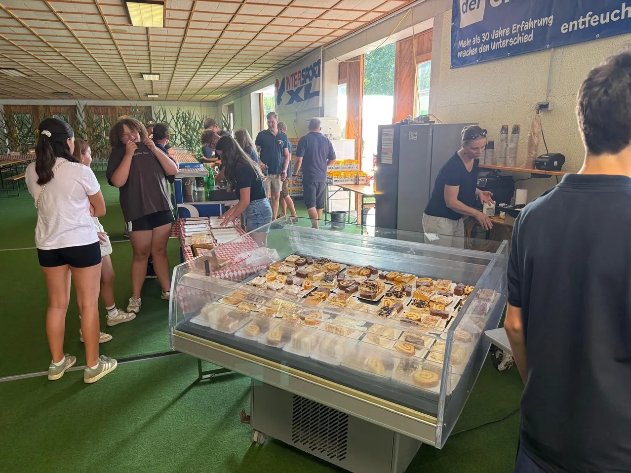 Eine Gruppe von Menschen versammelt sich um eine Glasvitrine mit verschiedenen Desserts in einem Raum mit grünem Teppich und einem Banner an der Wand.
