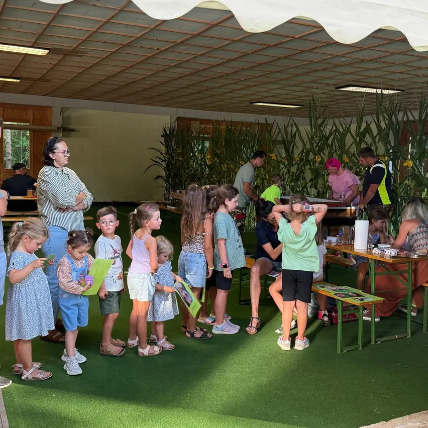 Eine Gruppe von Kindern und Erwachsenen steht in einem Raum mit grünem Teppich und Tischen, einige schauen sich Gegenstände auf den Tischen an.