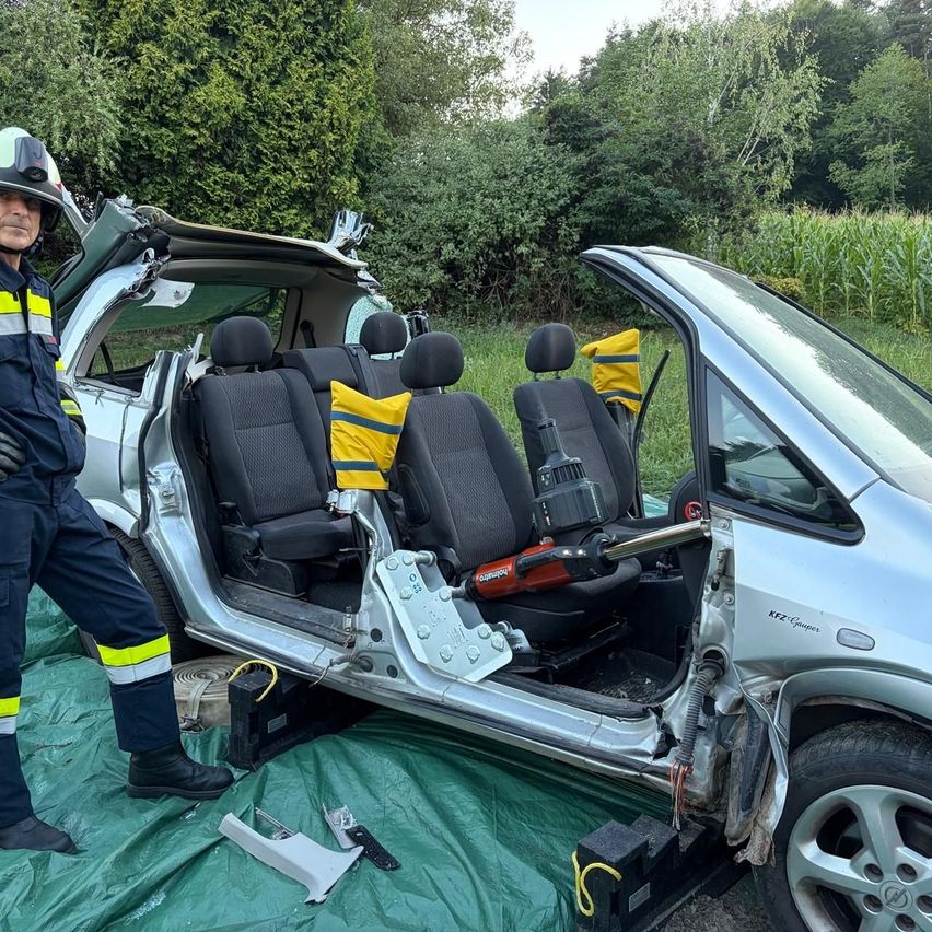 Ein Feuerwehrmann steht neben einem beschädigten Auto mit offenen Türen, das Innere mit Sitzen und Werkzeugen auf dem Boden zeigt.