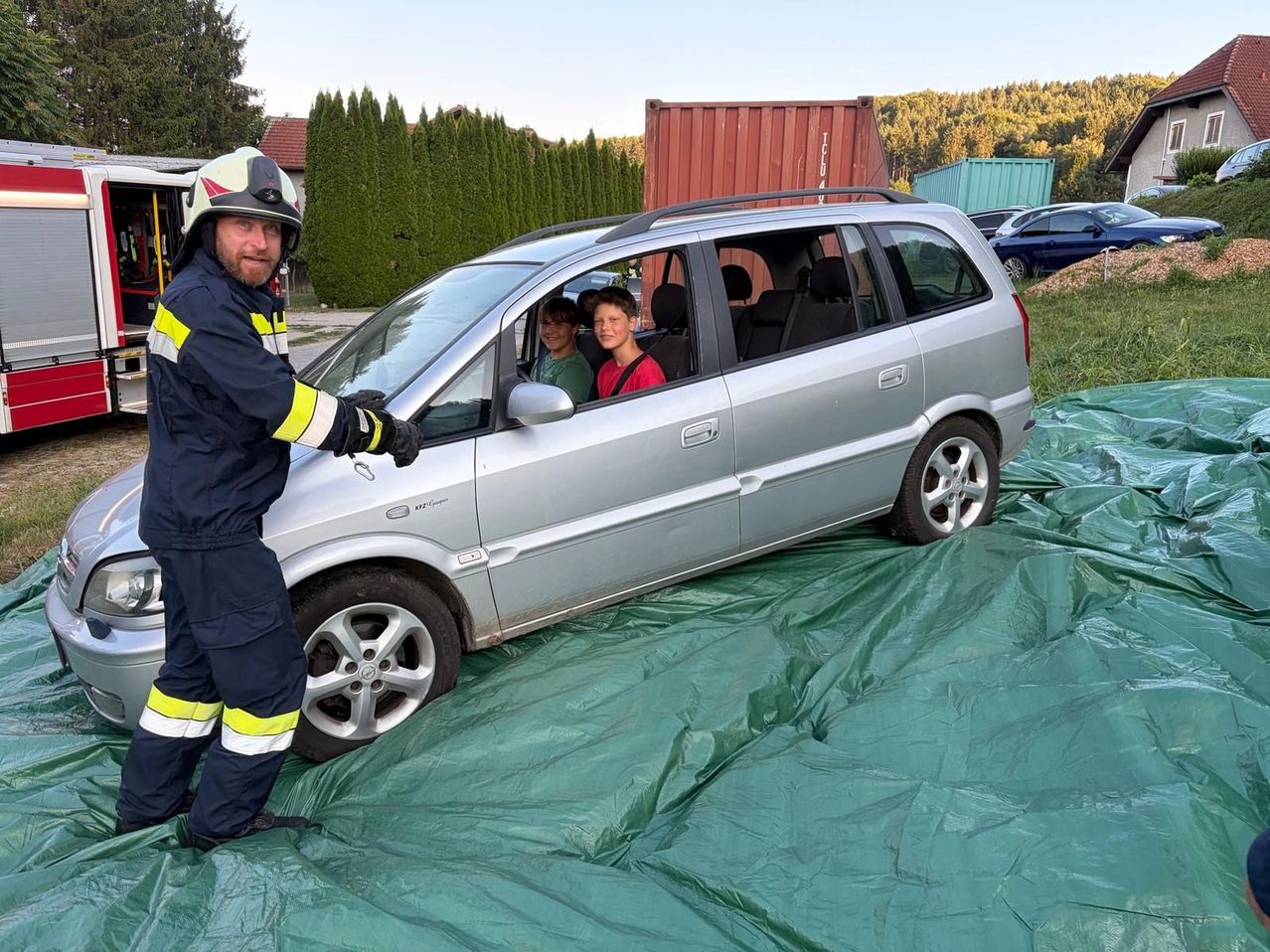 Ein Feuerwehrmann in voller Ausrüstung steht neben einem silbernen Auto mit zwei Jungen im Inneren auf einer grünen Plane. Hinter dem Auto steht ein roter Container und ein blaues Auto, das auf Gras geparkt ist.