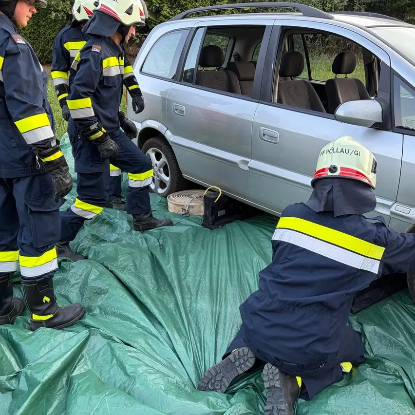 Eine Gruppe von Feuerwehrleuten in Schutzkleidung arbeitet an einem silbernen Auto, einer von ihnen kniet und scheint einen Reifen zu wechseln.