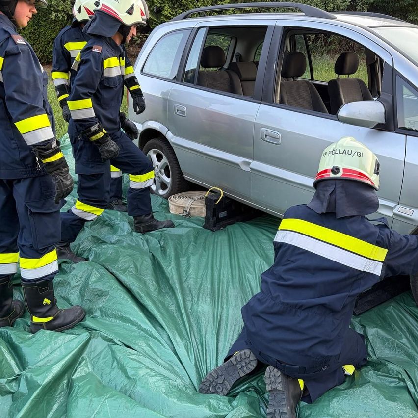 Eine Gruppe von Feuerwehrleuten in Schutzkleidung arbeitet an einem silbernen Auto, einer von ihnen kniet und scheint einen Reifen zu wechseln.