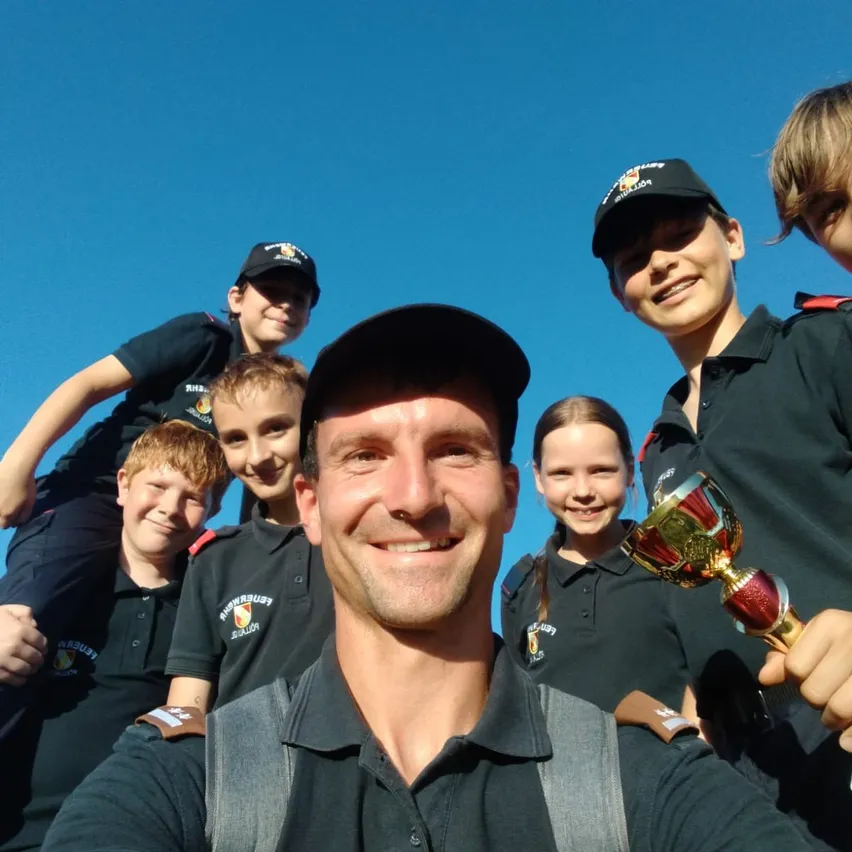 Eine Gruppe lächelnder Kinder und ein Mann in schwarzen Hemden und Mützen. Der Mann hält eine Trophäe. Die Kinder stehen hinter ihm. Sie sind draußen bei klarem blauem Himmel.