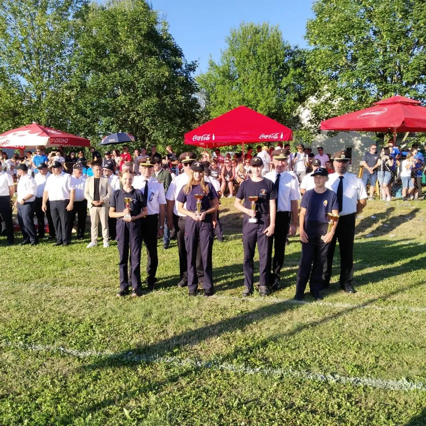 Eine Gruppe von Personen in Uniformen steht auf einem Rasenfeld mit roten Zelten im Hintergrund und hält Trophäen.