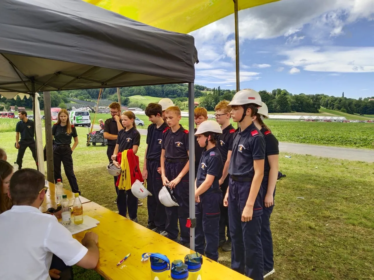 Eine Gruppe junger Menschen in Uniformen steht unter einem Zelt. Sie tragen weiße Helme und scheinen an einer Veranstaltung teilzunehmen. Dahinter ist ein saftiges Feld.