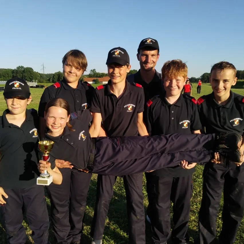 Eine Gruppe junger Leute in Uniformen mit Mützen und Schuhen posiert für ein Foto. Eine Mädchen in der Mitte hält einen Pokal. Dahinter ist ein verschwommener Hintergrund aus Gras und Bäumen.