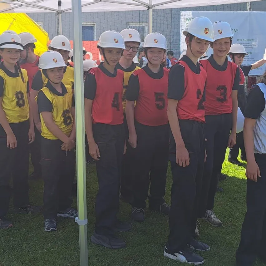 Eine Gruppe von Kindern in roten und gelben Trikots mit Nummern steht unter einem Zelt. Sie tragen weiße Helme und schwarze Hosen.