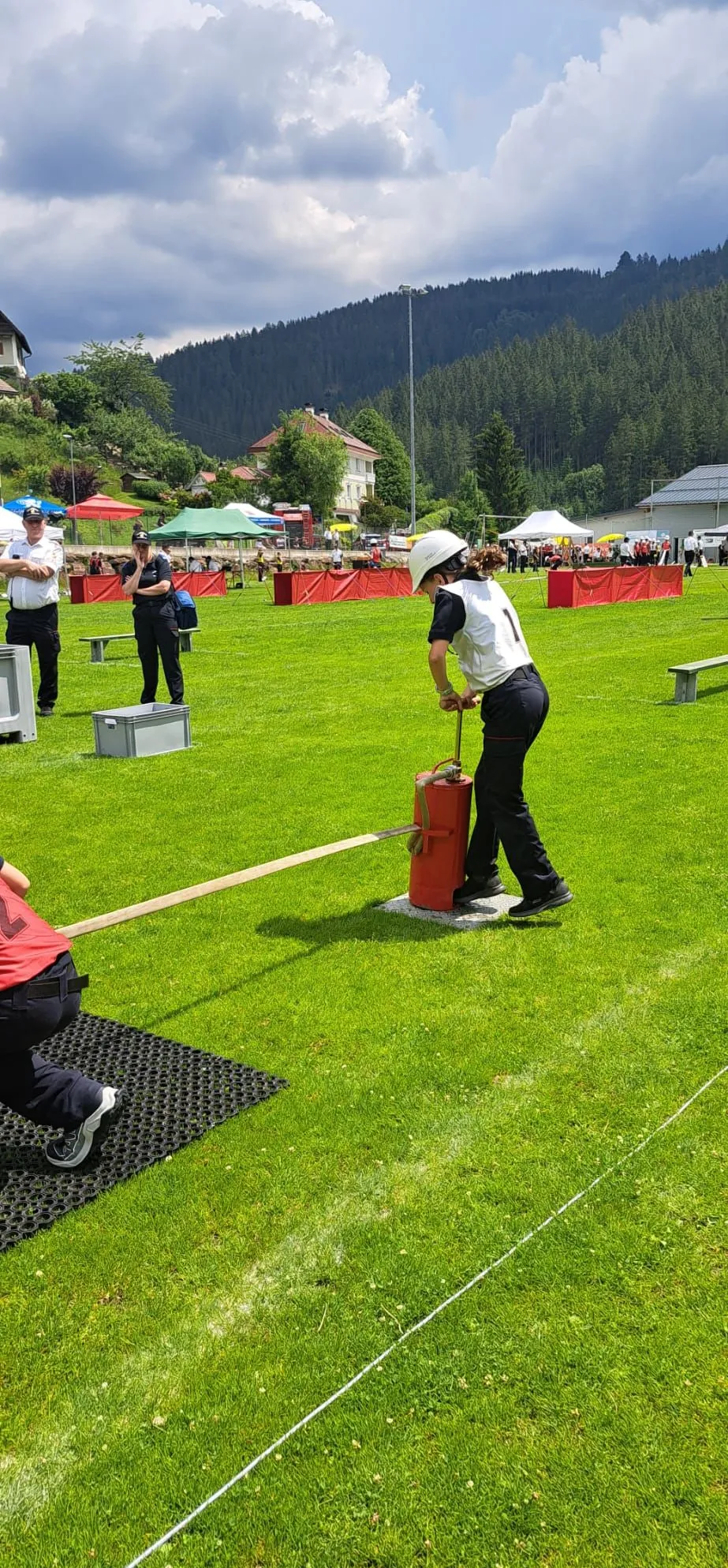 Eine Frau in einem weißen Helm und schwarzen Hosen zieht einen roten Feuerlöscher mit einem Seil. Eine andere Person ist am Boden und zieht das Seil.