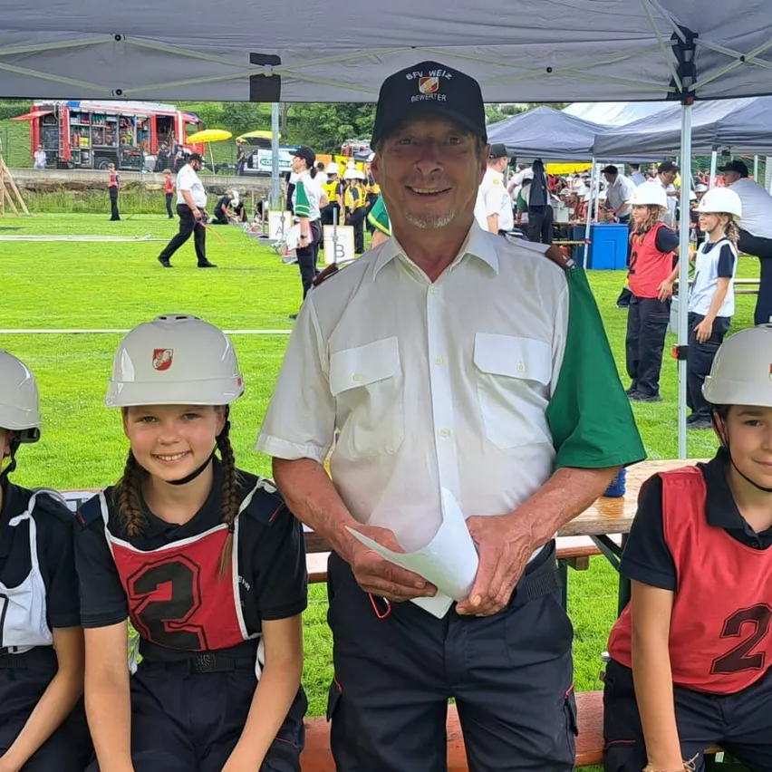 Ein älterer Mann steht unter einem Zelt mit drei Kindern, die alle Schutzhelme tragen. Der Mann hält ein Papier und lächelt. Im Hintergrund sind ein Feuerwehrauto und Personen zu sehen.