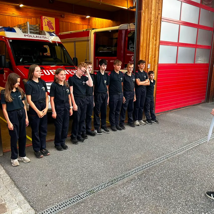 Eine Gruppe von Kindern in Uniform steht vor einer Feuerwache mit zwei Feuerwehrfahrzeugen darin.