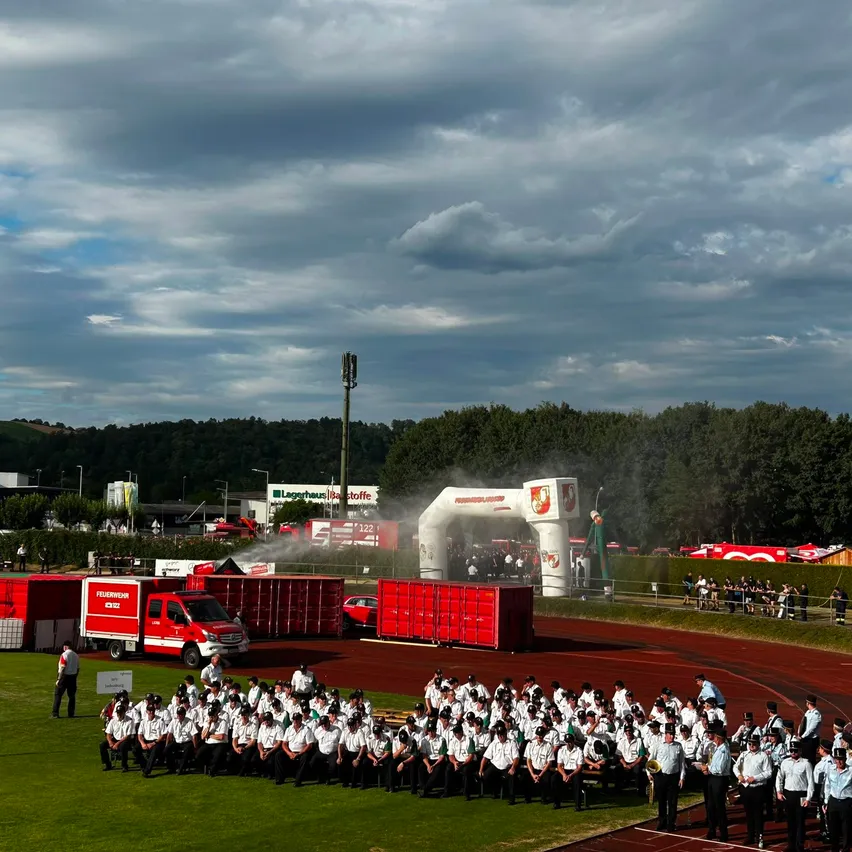 Eine Gruppe Feuerwehrleute posiert für ein Foto vor einem roten Fahrzeug und roten Containern auf einem Rasenfeld. Dahinter steht ein weißer Bogen. Bäume und ein bewölkter Himmel sind im Hintergrund.