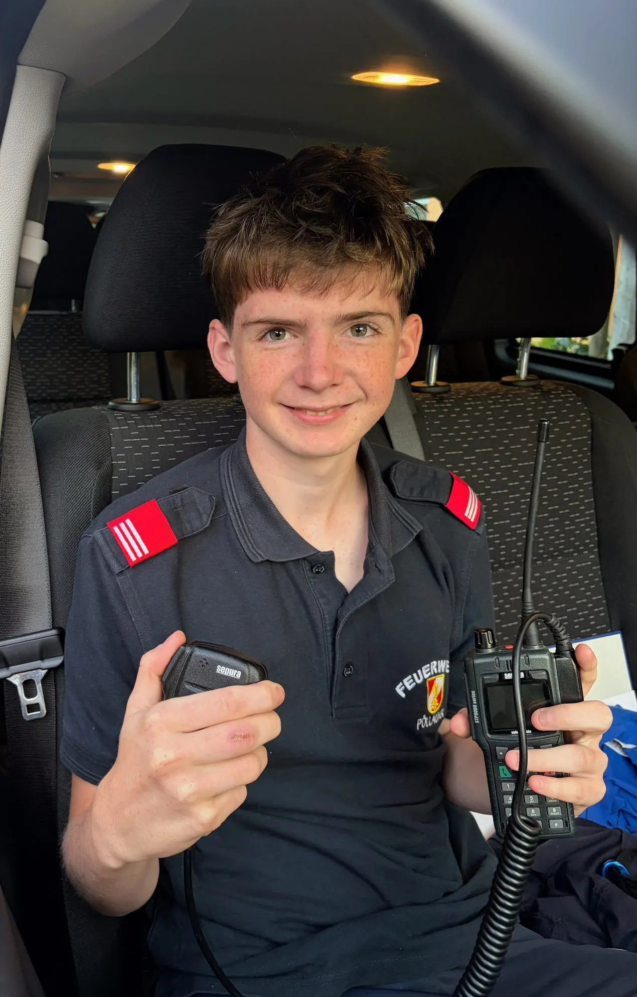 Ein junger Junge in einem schwarzen Shirt mit roten Aufnähern auf den Schultern sitzt auf dem Rücksitz eines Autos und hält zwei Walkie-Talkies.