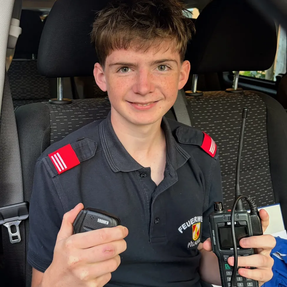 Ein junger Junge in einem schwarzen Shirt mit roten Aufnähern auf den Schultern sitzt auf dem Rücksitz eines Autos und hält zwei Walkie-Talkies.