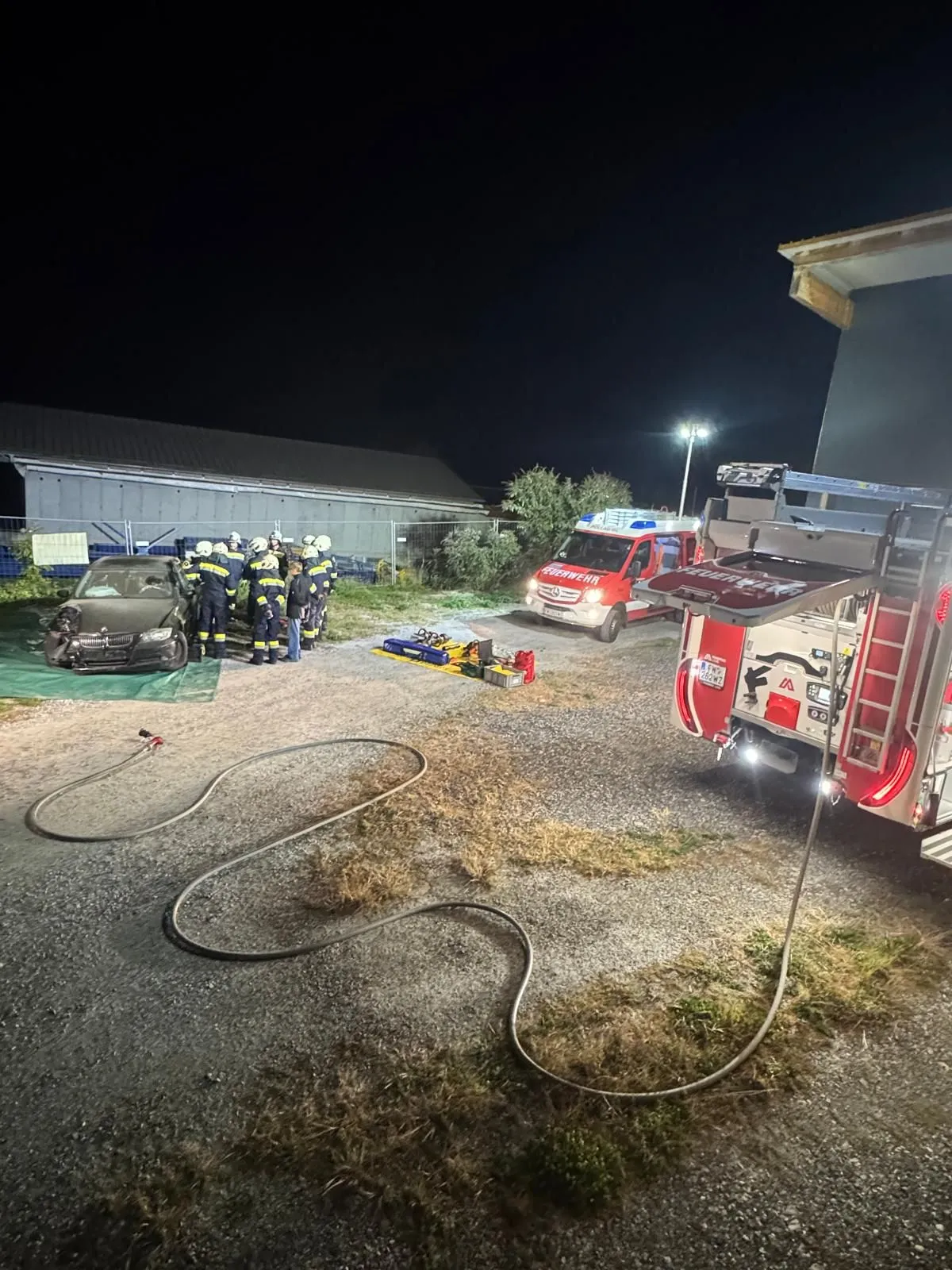 Eine Gruppe von Feuerwehrleuten versammelt sich um ein demoliertes Auto auf einem Parkplatz in der Nacht.