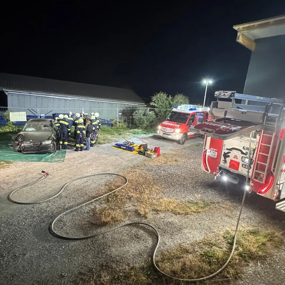 Eine Gruppe von Feuerwehrleuten versammelt sich um ein demoliertes Auto auf einem Parkplatz in der Nacht.