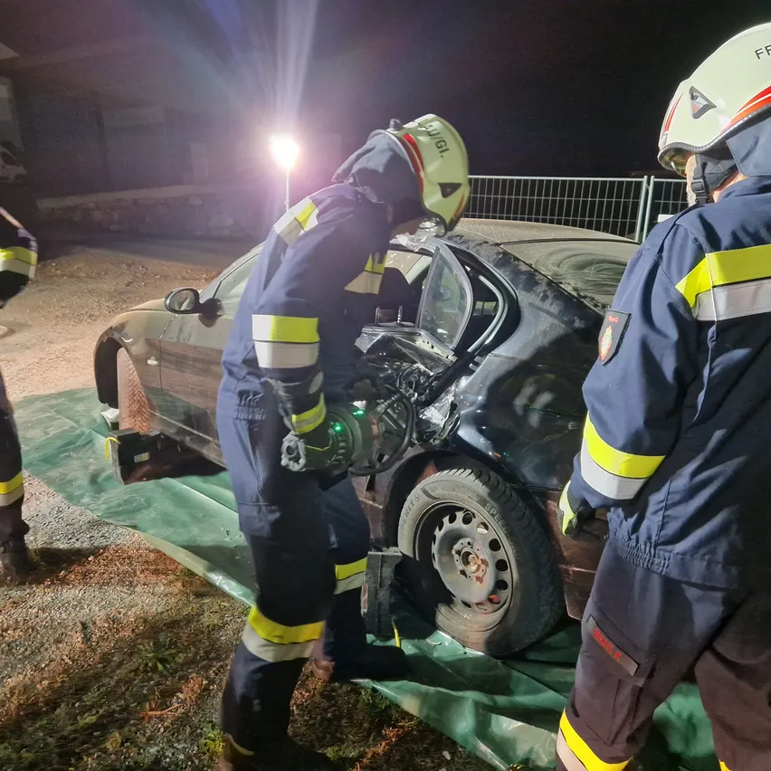 Drei Feuerwehrleute in Schutzkleidung arbeiten bei Nacht an einem beschädigten Auto. Einer benutzt ein Werkzeug am Rad des Autos, während die anderen daneben stehen.
