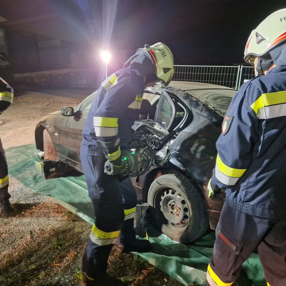 Drei Feuerwehrleute in Schutzkleidung arbeiten bei Nacht an einem beschädigten Auto. Einer benutzt ein Werkzeug am Rad des Autos, während die anderen daneben stehen.