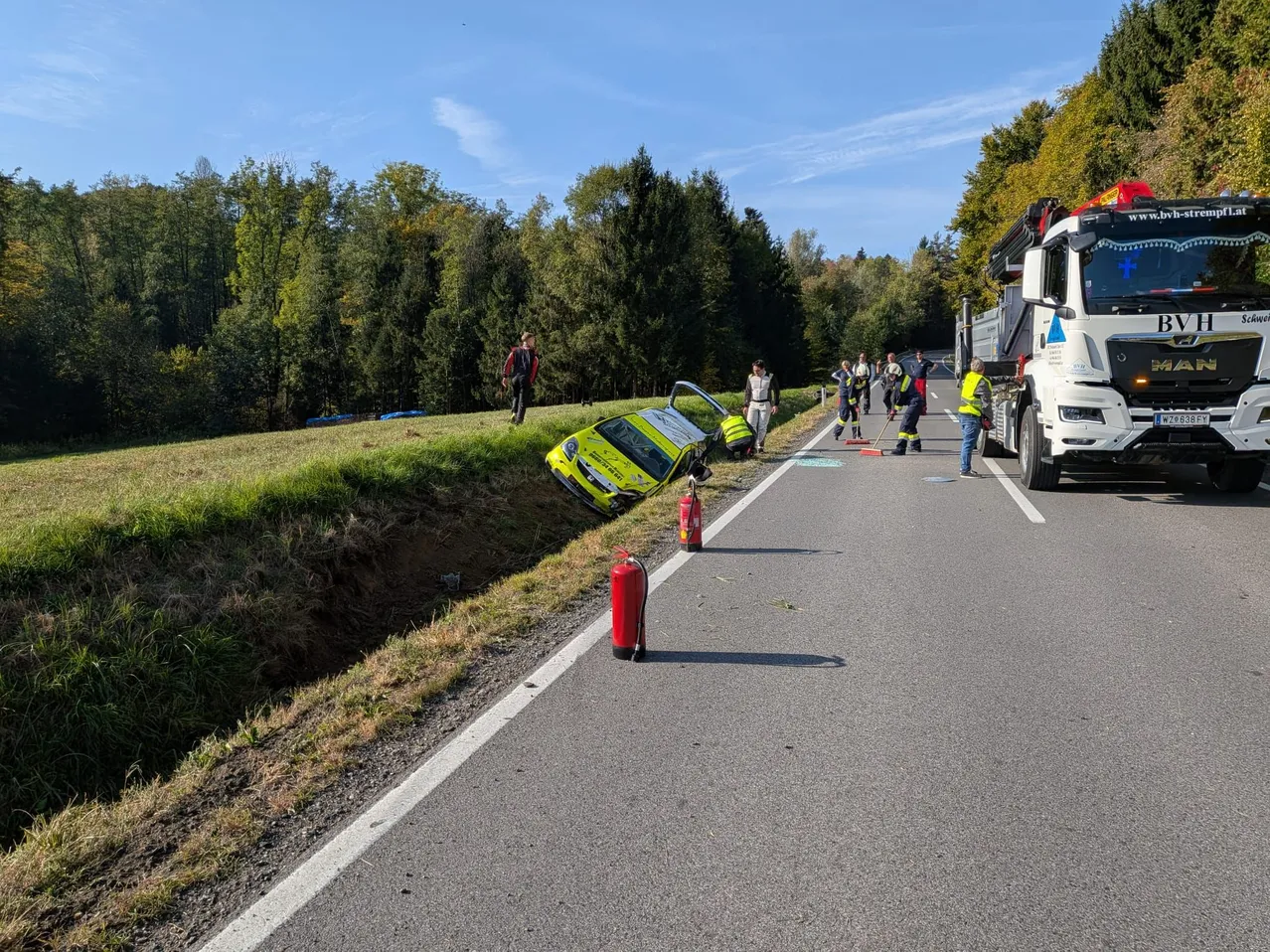Ein gelbes Auto ist auf der Straßenseite umgekippt. Ein Lastwagen ist auf der Seite geparkt, und mehrere Leute stehen darum herum.