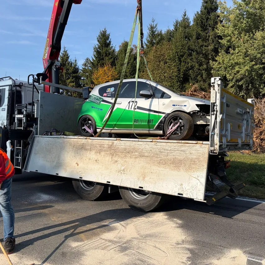 Ein Kran hebt ein beschädigtes grünes und weißes Auto auf einen Tieflader, der am Straßenrand steht.